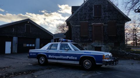 1991 Ford LTD Crown Victoria - Side view of a 1991 Ford Crown Victoria police car parked near old wooden buildings under a cloudy sky.