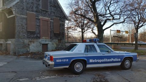 1991 Ford LTD Crown Victoria - A 1991 Ford Crown Victoria police car is parked next to a derelict building, its 'Metropolitan Police' markings visible.