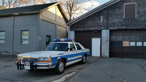 1991 Ford LTD Crown Victoria - A 1991 Ford Crown Victoria police car is parked outside a weathered garage with a large overhead door.