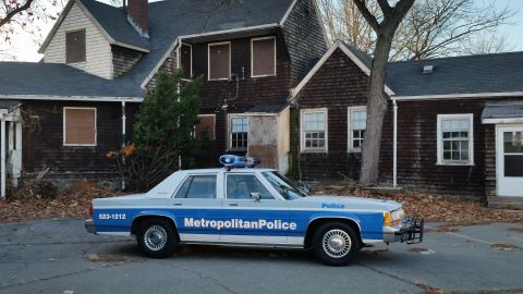1991 Ford LTD Crown Victoria - A 1991 Ford Crown Victoria police car is parked on a street in front of a dilapidated house.