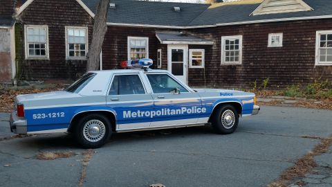 1991 Ford LTD Crown Victoria - Side view of a 1991 Ford Crown Victoria police car parked in front of a weathered building.