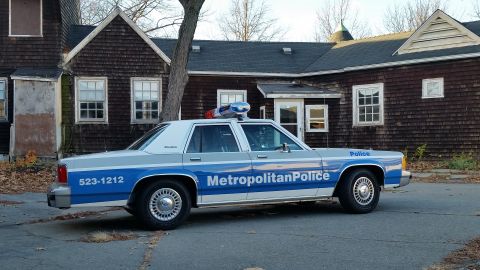 1991 Ford LTD Crown Victoria - A police car with 'Metropolitan Police' and a phone number is parked outside an old, shingled building.