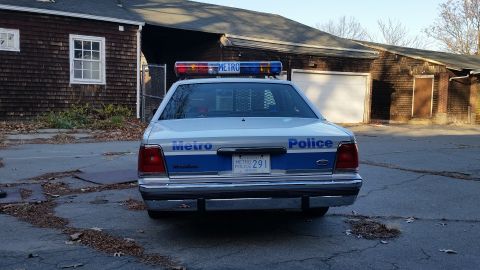 1991 Ford LTD Crown Victoria - A police car with a light bar is parked in front of a weathered building with a garage door.