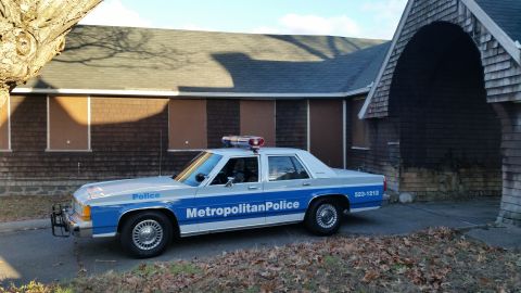 1991 Ford LTD Crown Victoria - A police car with a light bar sits parked in front of a building with boarded-up windows.