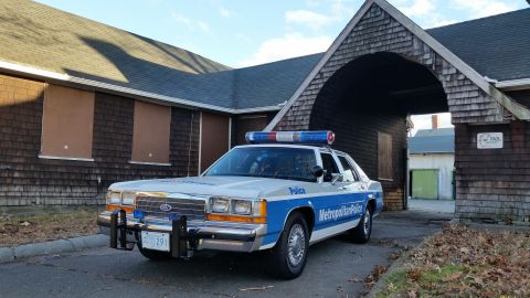 1991 Ford LTD Crown Victoria - A police car with a light bar is parked in front of an old building.