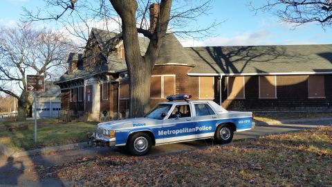 1991 Ford LTD Crown Victoria - A police car is parked on a street in front of a large, boarded-up building on a sunny day.