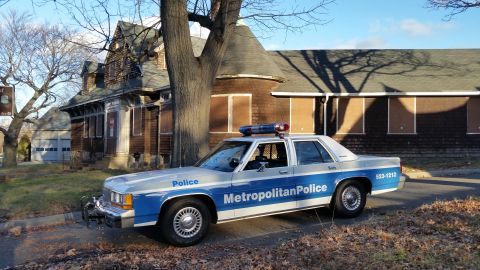 1991 Ford LTD Crown Victoria - A police car with 'Metropolitan Police' written on its side is parked in front of an old, boarded-up house.