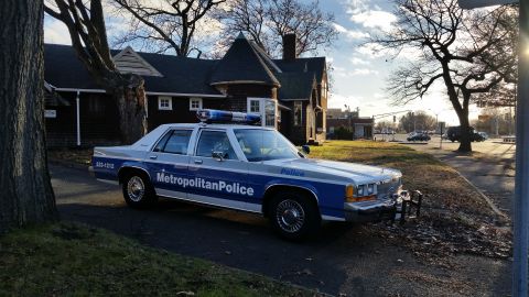 1991 Ford LTD Crown Victoria - A police car with a light bar is parked on a street next to a house on a sunny day.