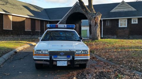 1991 Ford LTD Crown Victoria - A 1991 Ford Crown Victoria police car is parked in front of an old building with boarded-up windows.