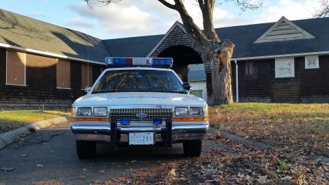 1991 Ford LTD Crown Victoria - A 1991 Ford Crown Victoria police car is parked in front of a weathered building with boarded-up windows.