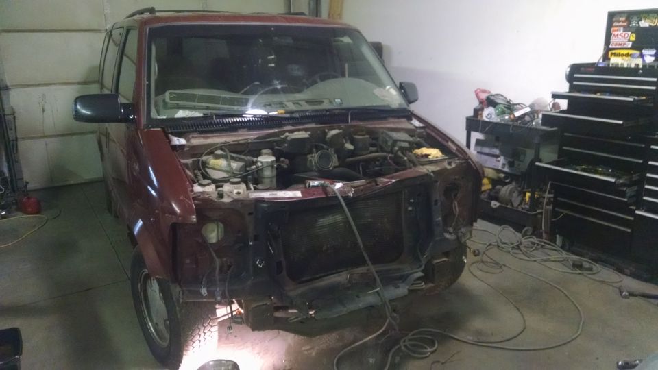 Maroon Chevrolet Astro van with front end removed, showing engine and radiator area. Tool chests and equipment are in the background