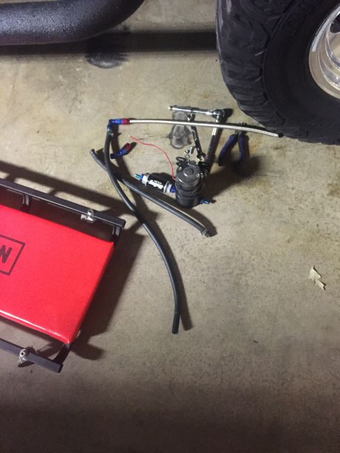 1982 Ford Bronco - A fuel pump assembly with braided lines and a wrench are laid out on a concrete floor, next to a red creeper and a vehicle tire.
