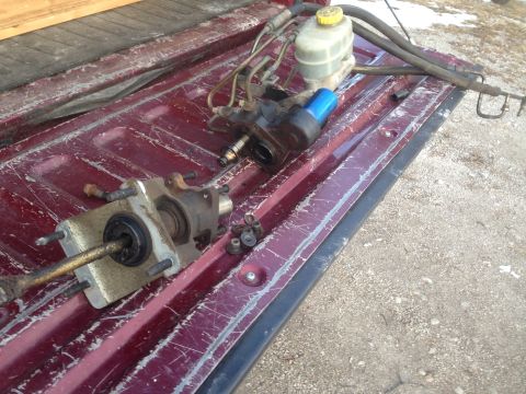 1979 Ford Bronco - A brake master cylinder and reservoir assembly are laid out on the bed of a dark red pickup truck. The parts appear to be used and are rest…