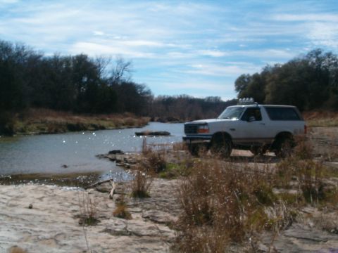 1996 Ford Bronco - A white Ford Bronco is parked on a rocky bank next to a flowing river under a cloudy sky.