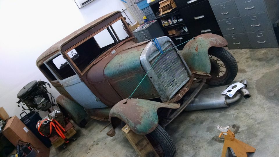 Rusty 1929 Ford Sedan project car in a garage with engine and exhaust parts nearby.
