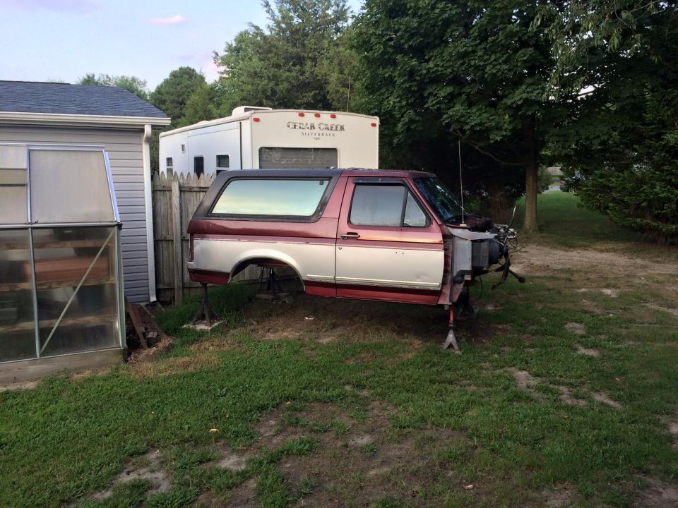 1994 Ford Bronco body on jack stands in a yard with a greenhouse and RV.