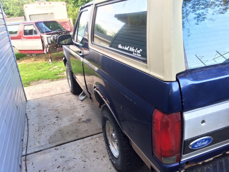 Rear driver's side of a dark blue Ford Bronco parked on a concrete driveway. Red and white pickup in background.