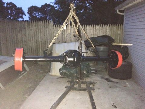 1994 Ford Bronco - A black rear axle assembly with bright orange brake drums is suspended by chains from an engine hoist. The assembly is outdoors in what app…