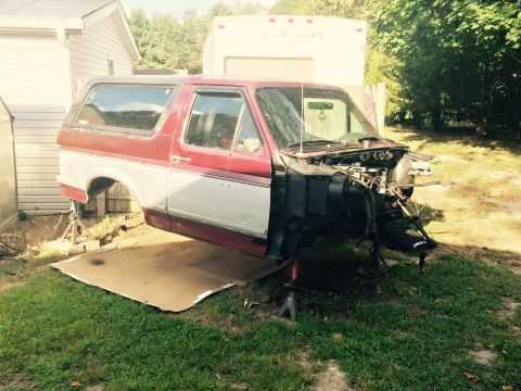 1994 Ford Bronco - A red and white Ford Bronco is disassembled and supported by jack stands in a grassy yard. The front end of the vehicle is missing, exposin…