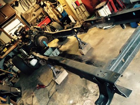 1994 Ford Bronco - A vehicle frame and engine are disassembled and supported by jack stands in a garage setting. Various tools and parts are scattered around…