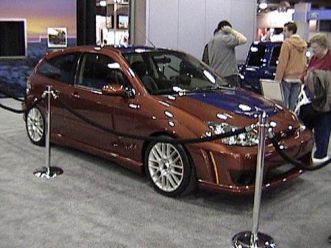 1995 Ford Bronco - An orange hatchback with a blue hood and custom body kit is shown at an auto show, with other vehicles and people in the background.