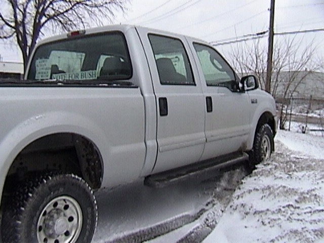 Silver Ford F-250 crew cab pickup, passenger side view, parked in snow with signs in rear window