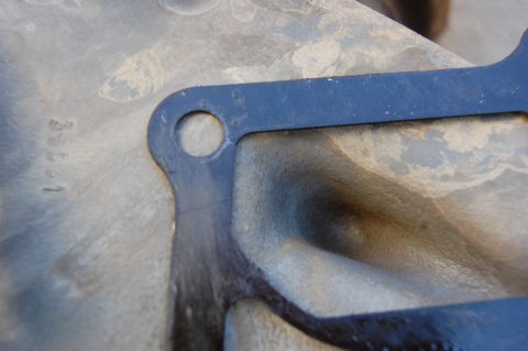 1994 Ford Bronco - A dark blue gasket is placed on a metal surface with visible casting marks and some dirt. The gasket has a hole in it and follows the conto…