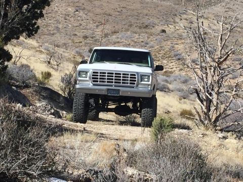 1979 Ford Bronco - A lifted white 1979 Ford Bronco is parked on a dirt road in a dry, hilly, desert landscape. The vehicle is viewed from a low angle, showing…