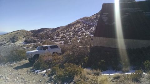 1979 Ford Bronco - A white 1970s Ford Bronco is parked on a dirt road next to a hillside with scattered snow. A large, dark structure with a sloped roof is to…