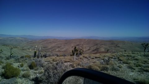1979 Ford Bronco - A wide-angle view of a desert landscape with sparse vegetation and rolling hills under a clear blue sky. The foreground shows a portion of…
