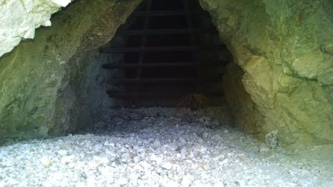 1979 Ford Bronco - The entrance to a dark cave or mine shaft is blocked by a metal grate. The ground in front of the opening is covered in rubble and rocks.