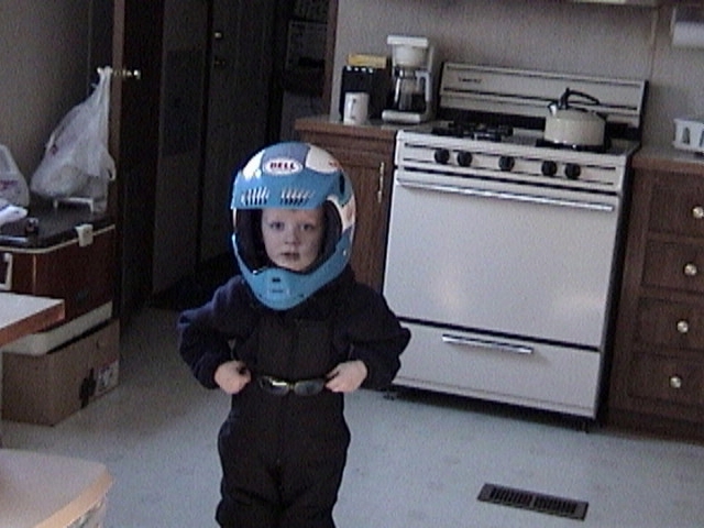 Small child in a kitchen wearing a dark racing suit and oversized blue Bell full-face helmet
