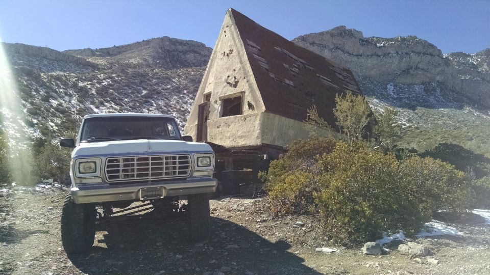 1979 Ford Bronco parked on dirt road in front of an abandoned A-frame cabin with mountains in the background.