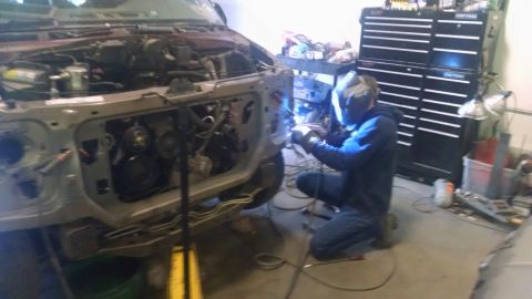 2000 Chevrolet Astro - A person wearing a welding mask and gloves is welding the front end of a vehicle. The vehicle's hood is open, revealing the engine compartm…