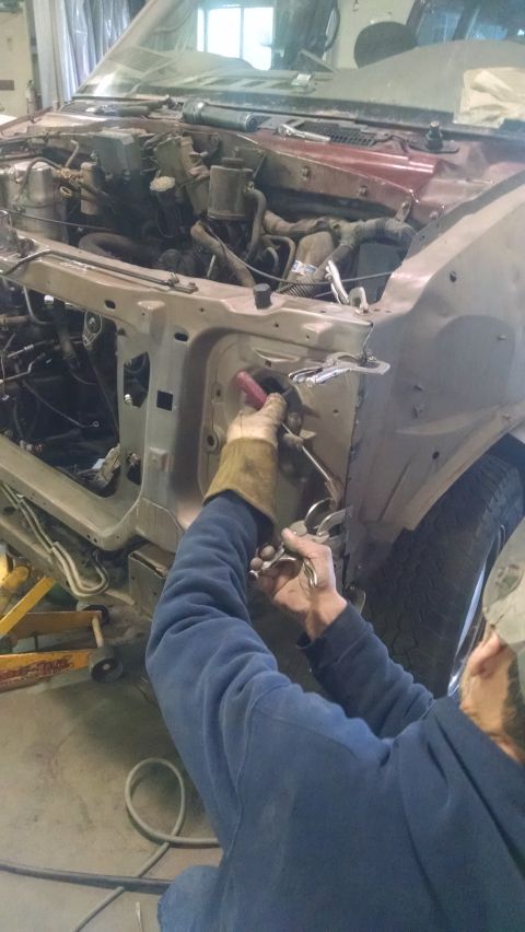 2000 Chevrolet Astro - Mechanic working with a tool inside a stripped engine bay and front core support of a vehicle in a garage.