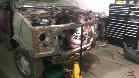2000 Chevrolet Astro - The front end of a maroon and silver van is shown in a workshop setting, with the hood open and engine exposed. Tools and a large black too…