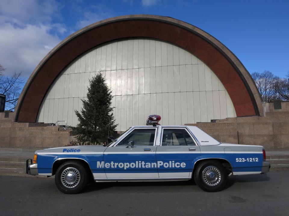 Blue and white 1991 Ford Crown Victoria police car parked outdoors with a large arch structure behind it