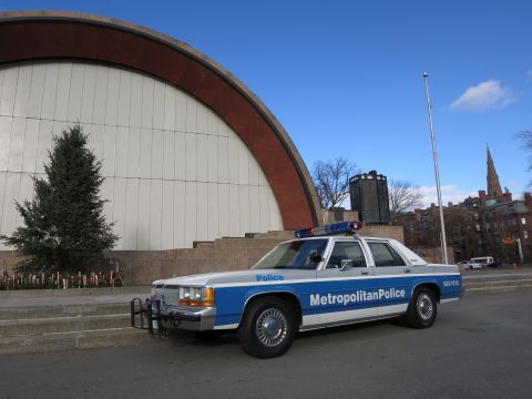 1991 Ford LTD Crown Victoria - A police car with emergency lights on top is parked in front of a large, curved building.