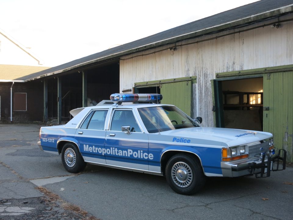 Blue and white Metropolitan Police Crown Victoria beside a weathered green barn