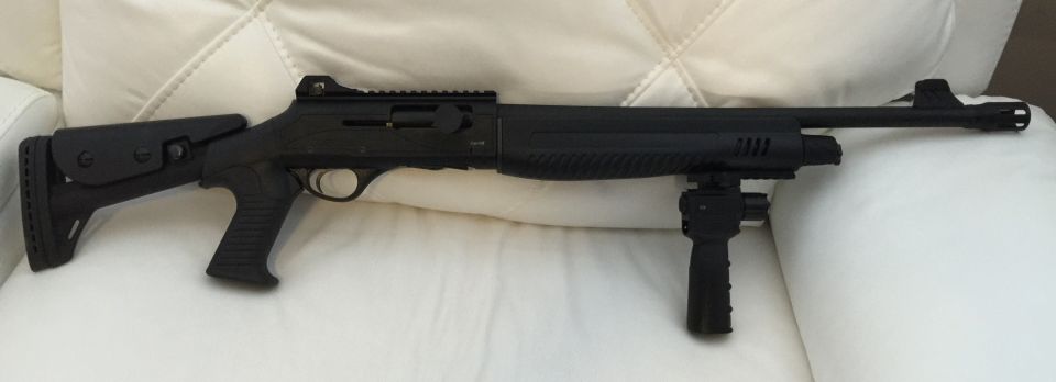 Black tactical shotgun with adjustable stock and vertical foregrip on a white couch.