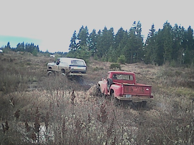 Red pickup truck and a white SUV driving through a muddy field with trees in the background