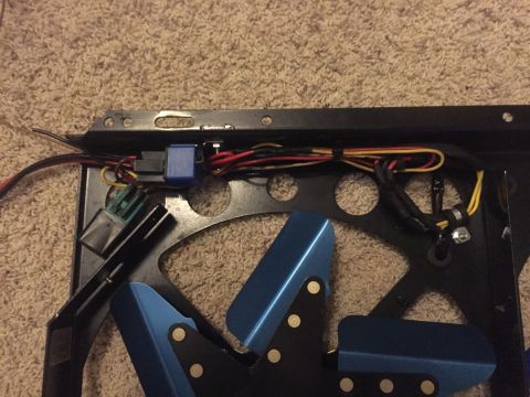 1982 Ford Bronco - A close-up view of wiring and electrical components attached to a black metal bracket, possibly for a fan, with blue fan blades visible bel…
