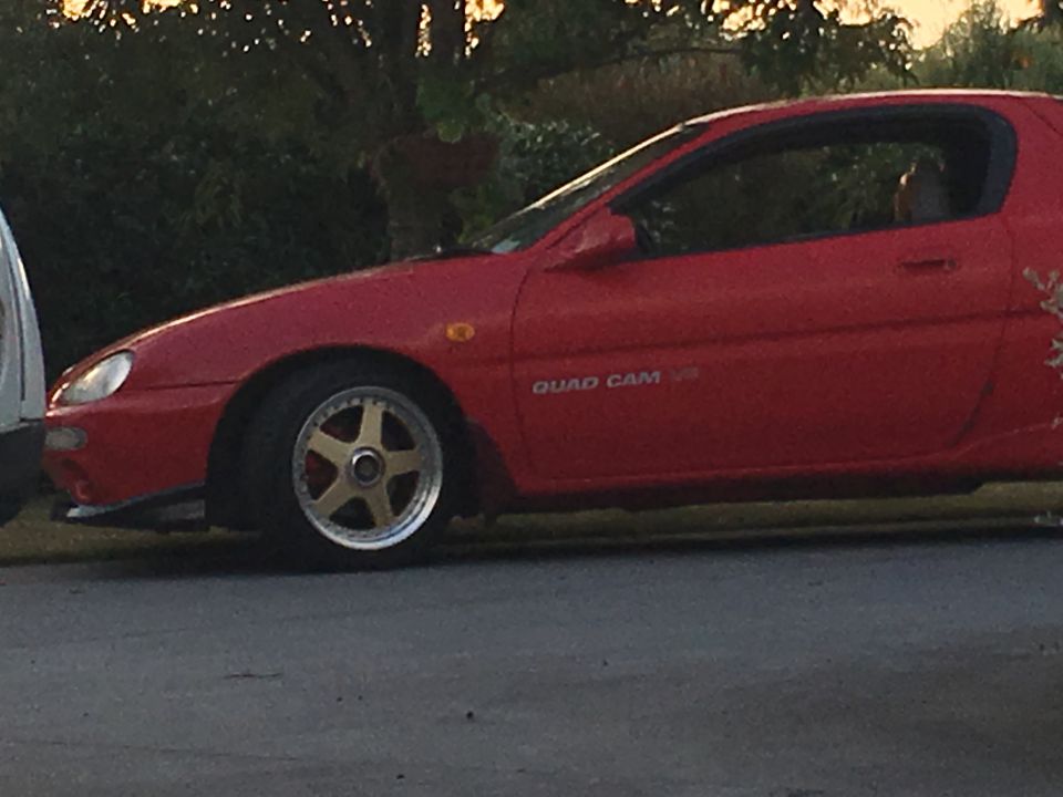 Red 1993 Mazda MX-3 coupe side view with gold wheels and Quad Cam V6 badge