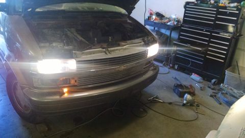 2000 Chevrolet Astro - The front of a Chevrolet Astro van with its hood open is parked in a garage. The headlights are on, illuminating the front grille and engin…