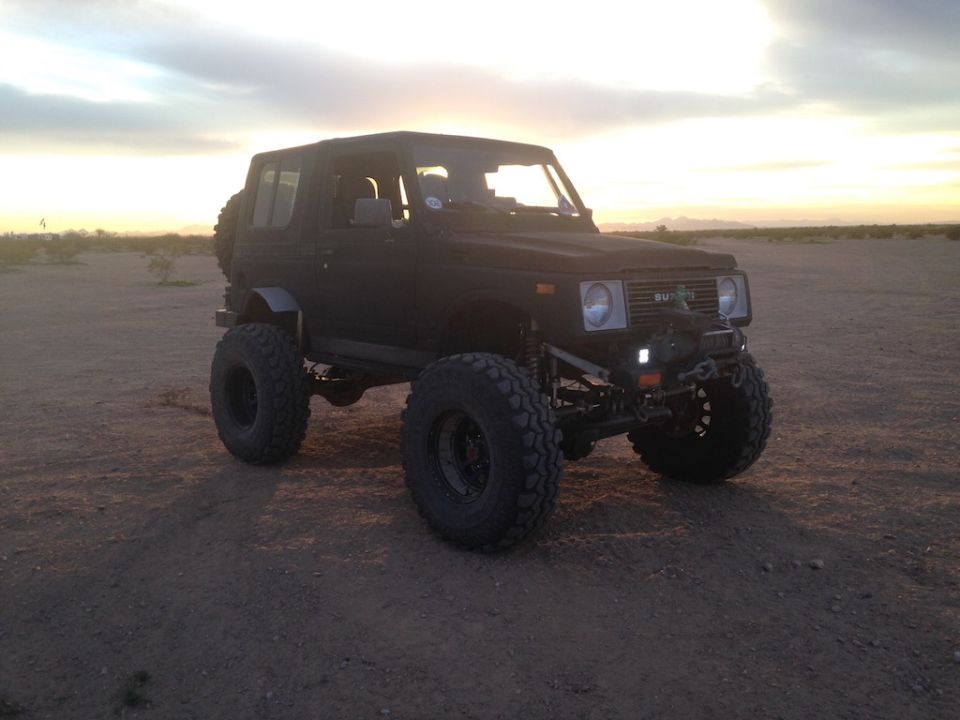 Black 1987 Suzuki Samurai with oversized tires and lifted suspension parked in a desert landscape at sunset