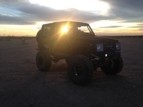 1987 Suzuki Samurai - A black 1987 Suzuki Samurai is shown off-road during a desert sunset, its large tires and lifted suspension ready for adventure.
