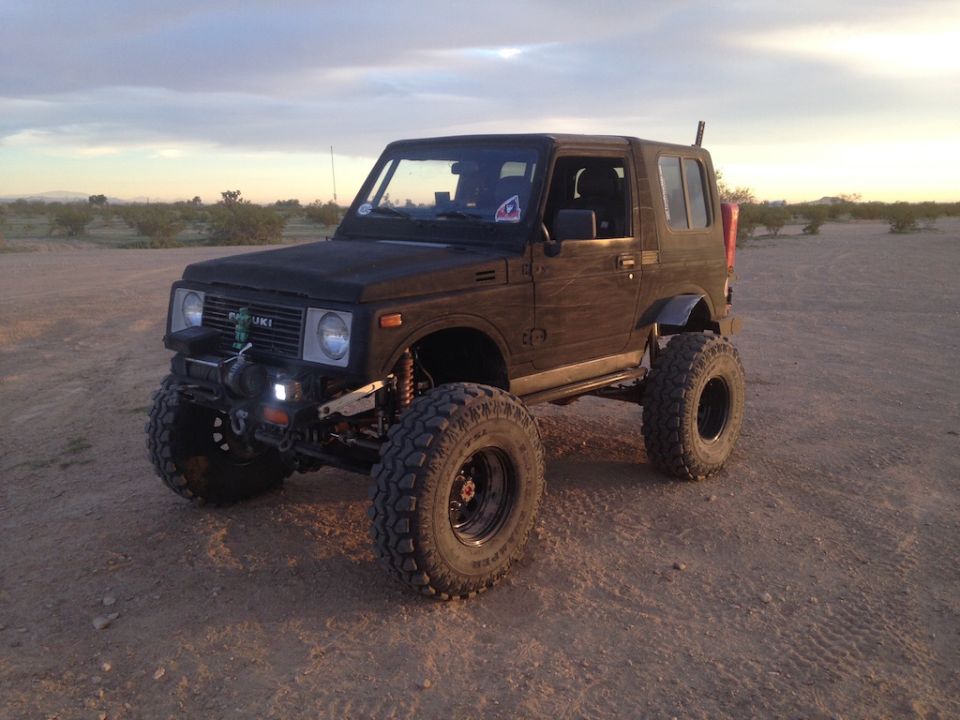 Black Suzuki Samurai with large tires and lifted suspension parked on dirt. Has a winch on the front