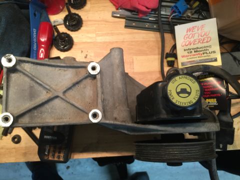 1996 Ford Bronco - Power steering pump and bracket assembly with pulley, on a workbench with tools and paperwork.
