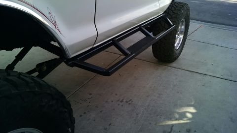 1979 Ford Bronco - A white SUV with custom side steps and large tires is parked on a concrete surface.