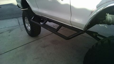1979 Ford Bronco - Aftermarket sliders are mounted to the side of a lifted white vehicle, with large off-road tires visible.